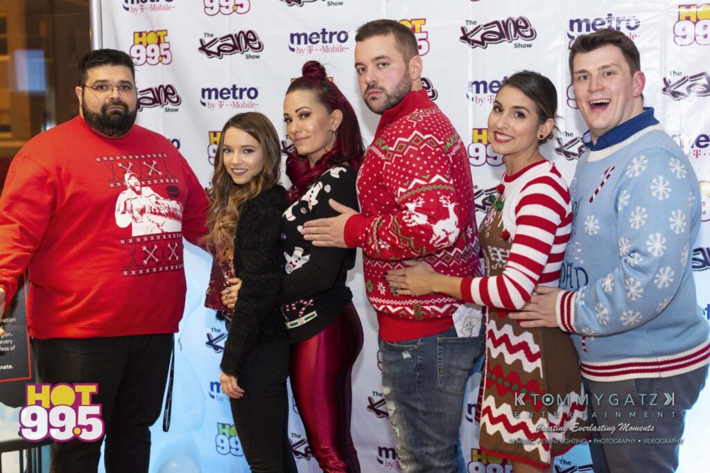Jingle-Ball-2018-BTS-03 - TOMMY GATZ ENTERTAINMENT A group of six people posing in festive holiday sweaters against a backdrop with logos.
