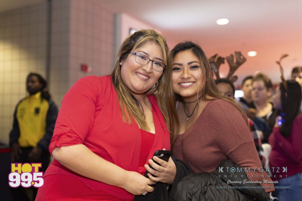 Jingle-Ball-2018-BTS-04 - TOMMY GATZ ENTERTAINMENT Two people smiling and posing together at an indoor event, with a crowd in the background. They are both wearing glasses and casual attire, with a logo visible in the corner of the image.