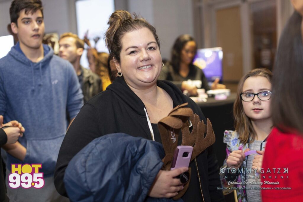 Jingle-Ball-2018-BTS-07 - TOMMY GATZ ENTERTAINMENT A woman holding a phone and reindeer antlers smiles at the camera. People are gathered in the background at an indoor event.