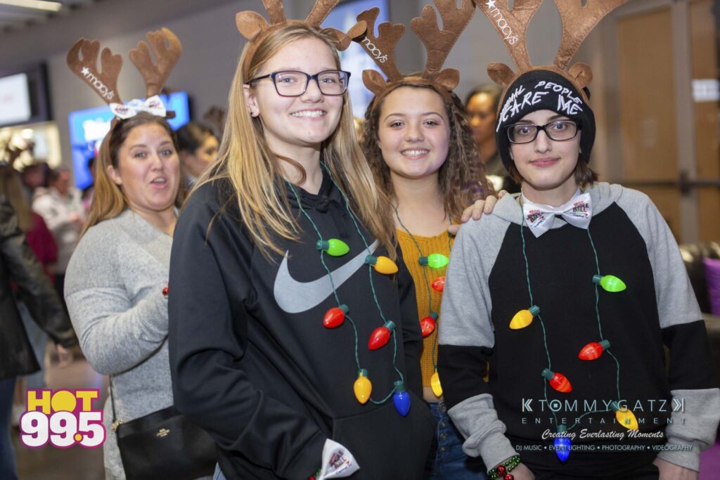 Jingle-Ball-2018-BTS-11 - TOMMY GATZ ENTERTAINMENT Group of people wearing reindeer antlers and holiday lights, one person has a hat with "Make Me" text. A "HOT 99.5" logo is in the corner.