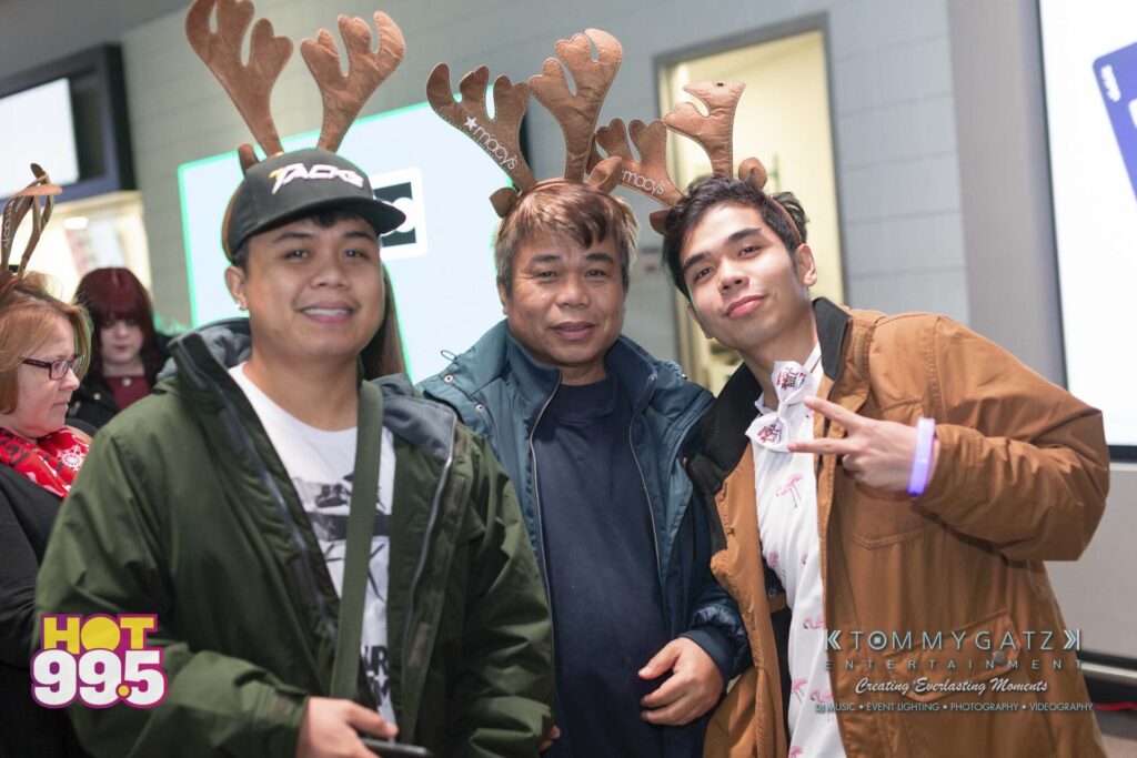 Jingle-Ball-2018-BTS-12 - TOMMY GATZ ENTERTAINMENT Three people wearing reindeer antlers pose for a photo indoors. One person makes a peace sign. The background shows a blurred crowd and a sign with "HOT 99.5" logo.