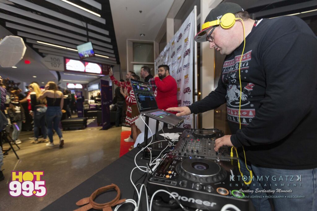 Jingle-Ball-2018-BTS-17 - TOMMY GATZ ENTERTAINMENT DJ in a festive sweater playing music on a console, wearing headphones. People in the background, some in holiday attire.