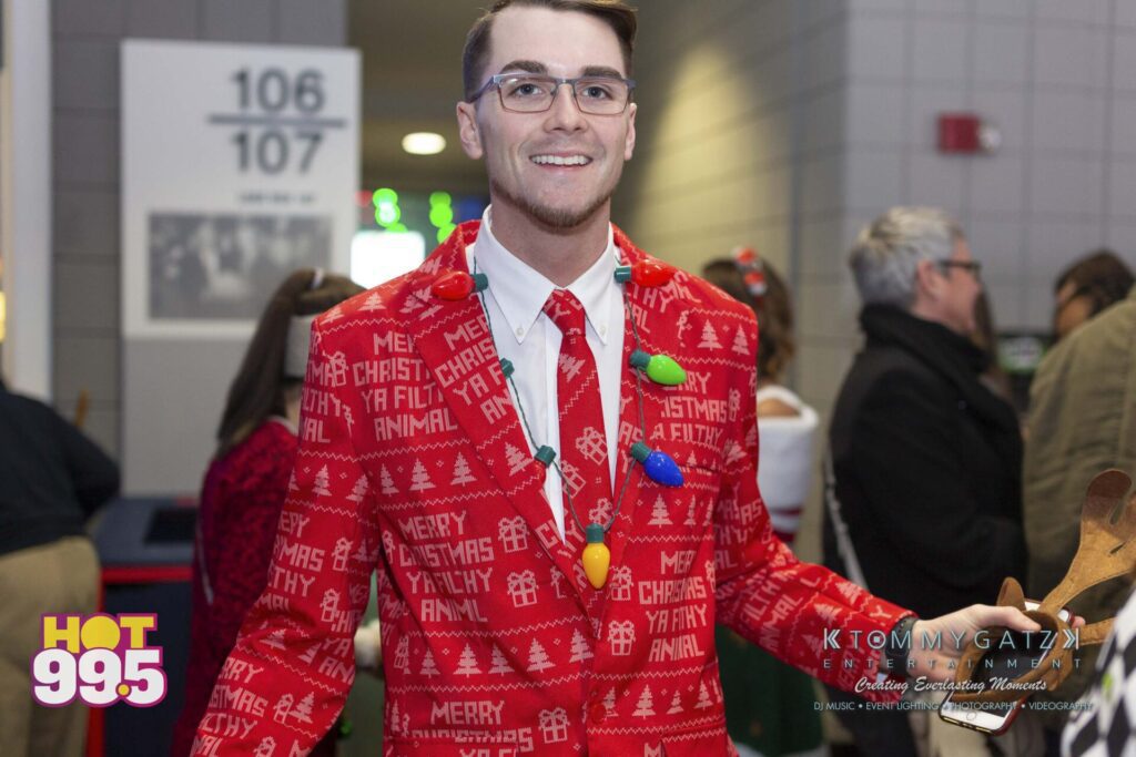Jingle-Ball-2018-BTS-19 - TOMMY GATZ ENTERTAINMENT Man in red holiday sweater with "Merry Christmas Ya Filthy Animal" text, holding a wooden item, standing indoors.