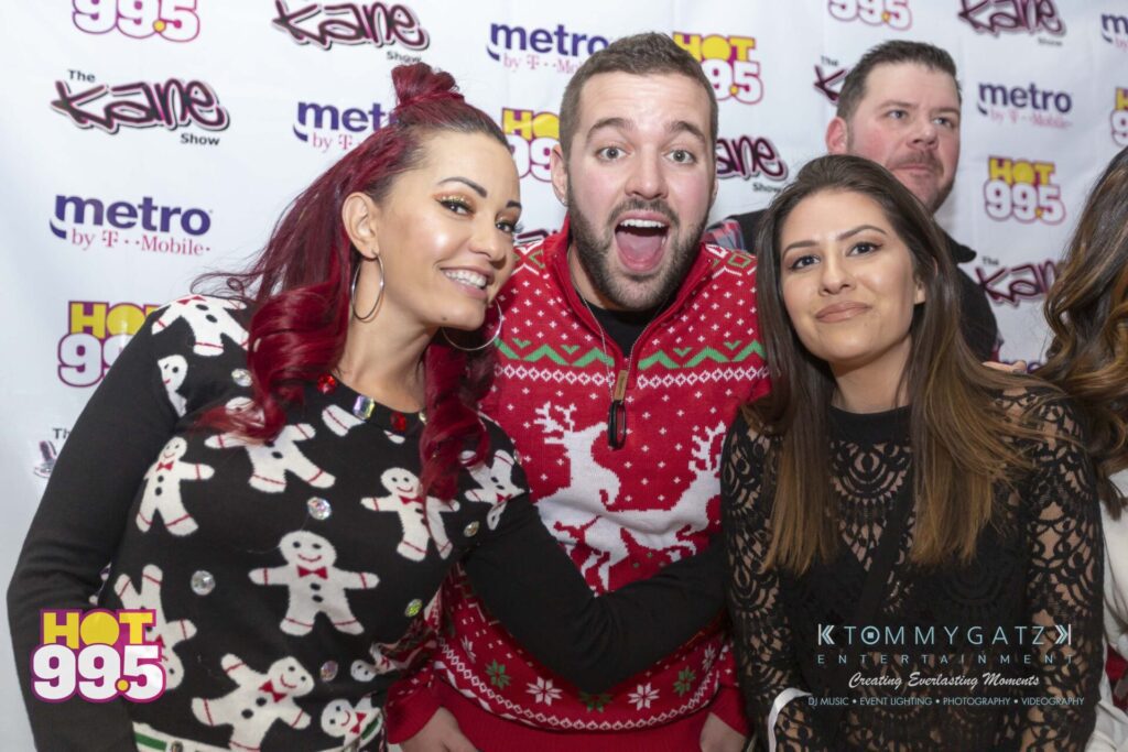 Jingle-Ball-2018-BTS-21 - TOMMY GATZ ENTERTAINMENT Three people smiling and posing in front of a branded backdrop featuring "HOT 99.5" and "The Kane Show" logos. One wears a festive sweater.