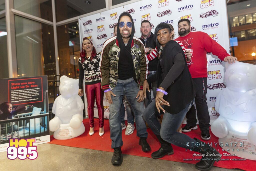 Jingle-Ball-2018-BTS-26 - TOMMY GATZ ENTERTAINMENT A group of people posing enthusiastically on a red carpet in front of a branded backdrop, with inflatable bear figures on either side.