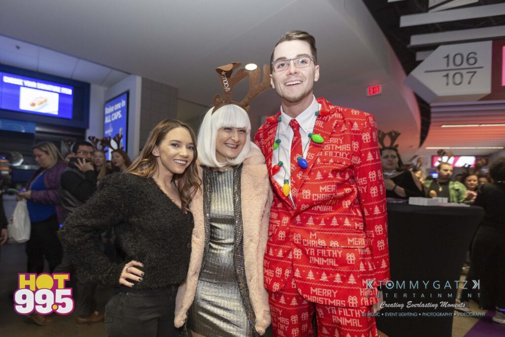 Jingle-Ball-2018-BTS-29 - TOMMY GATZ ENTERTAINMENT Three people dressed in festive holiday attire pose for a photo at an indoor event. Two wear reindeer antlers, and one is in a red Christmas suit. Signs for sections 106 and 107 are visible.