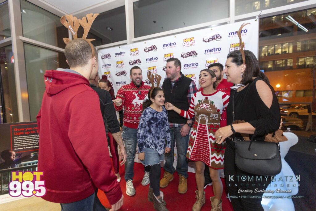 Jingle-Ball-2018-BTS-36 - TOMMY GATZ ENTERTAINMENT A group of people wearing festive attire stands in front of a backdrop with logos, including Hot 99.5. Some are wearing reindeer antlers.