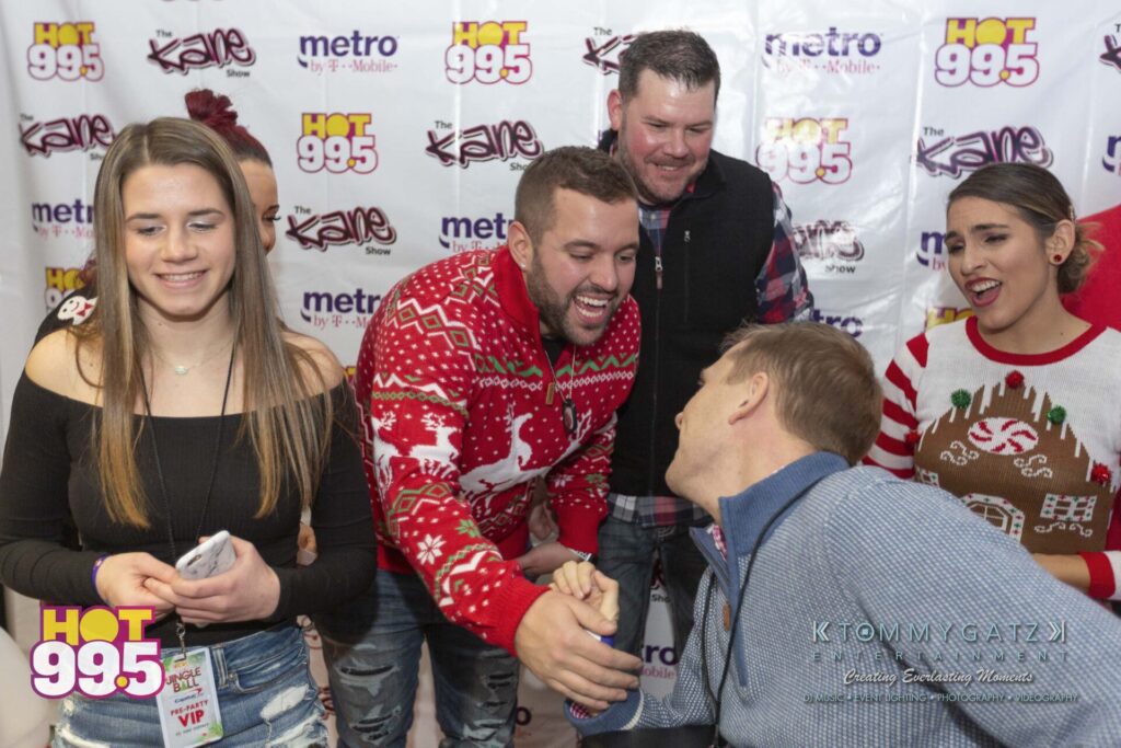 Jingle-Ball-2018-BTS-46 - TOMMY GATZ ENTERTAINMENT Group of people in festive sweaters at a Hot 99.5 event, with one person shaking hands.
