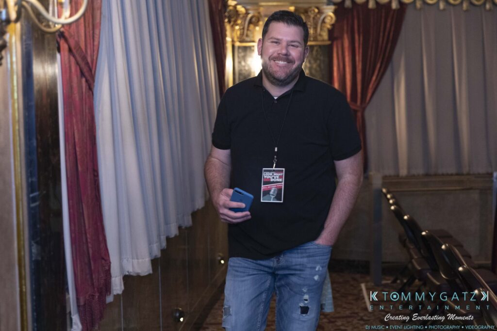 A man in casual clothing with a lanyard stands smiling in an ornate venue, holding a blue phone.