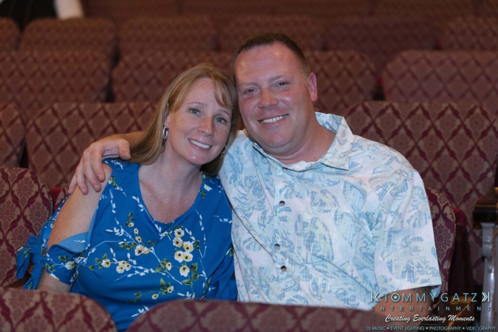 A man and woman sitting closely on patterned chairs in a theater. He is wearing a floral shirt, and she is in a blue dress with floral patterns. They are smiling at the camera.