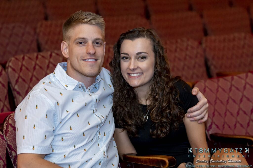 A smiling man and woman sit together in a theater with red patterned seats.