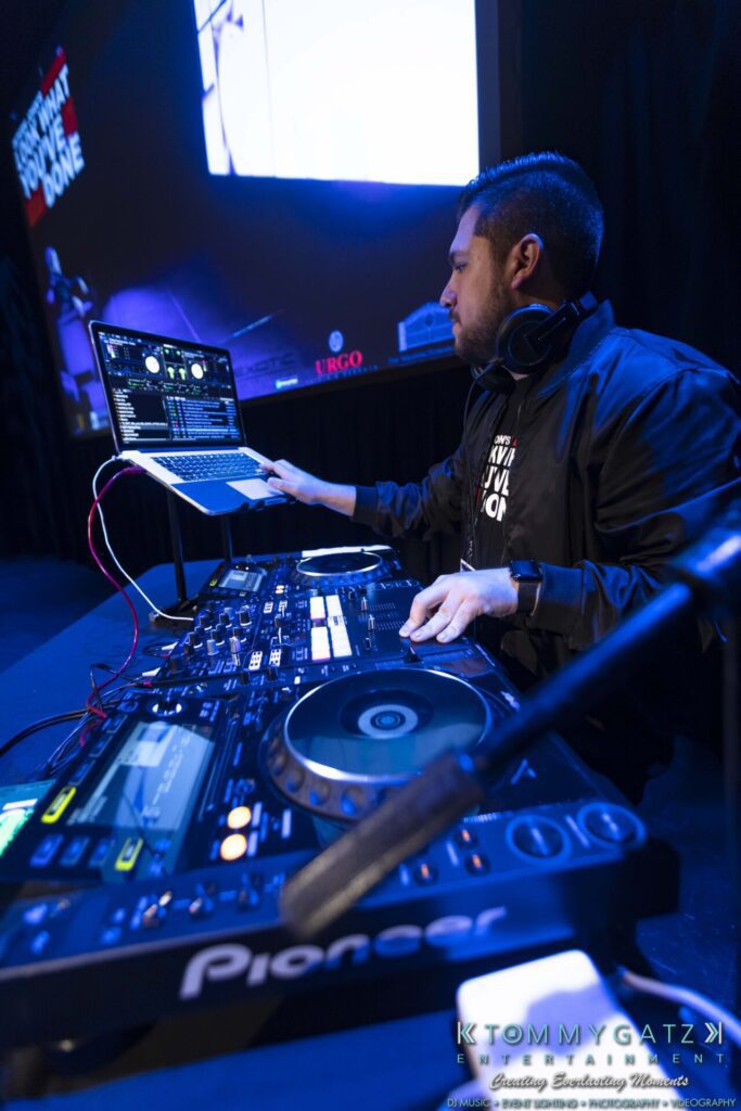 DJ performing on stage with equipment, including a laptop and turntable, under blue lighting. A projection screen is visible in the background.
