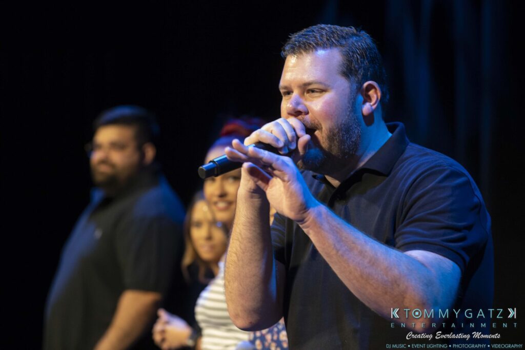 Man singing into a microphone on stage with a group of people in the background.