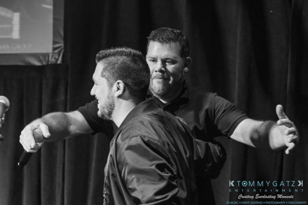 Two men in a black-and-white photo; one faces forward holding a microphone, and the other stands behind him with arms wide open, as if preparing for an embrace.