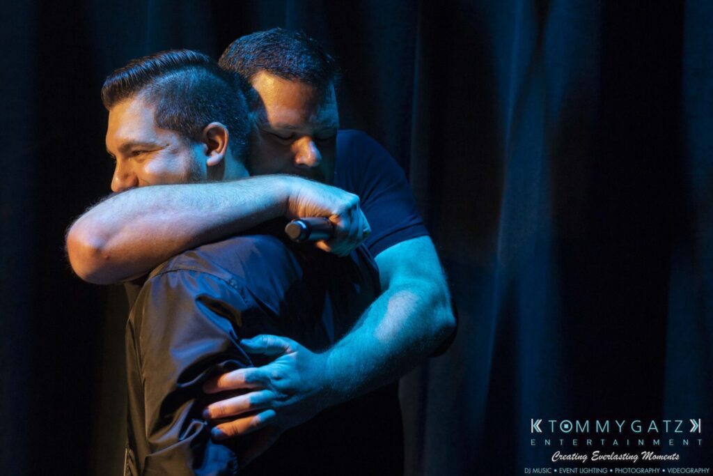 Two men embrace on stage, one holding a microphone. The background is a dark curtain and a logo is visible in the corner.