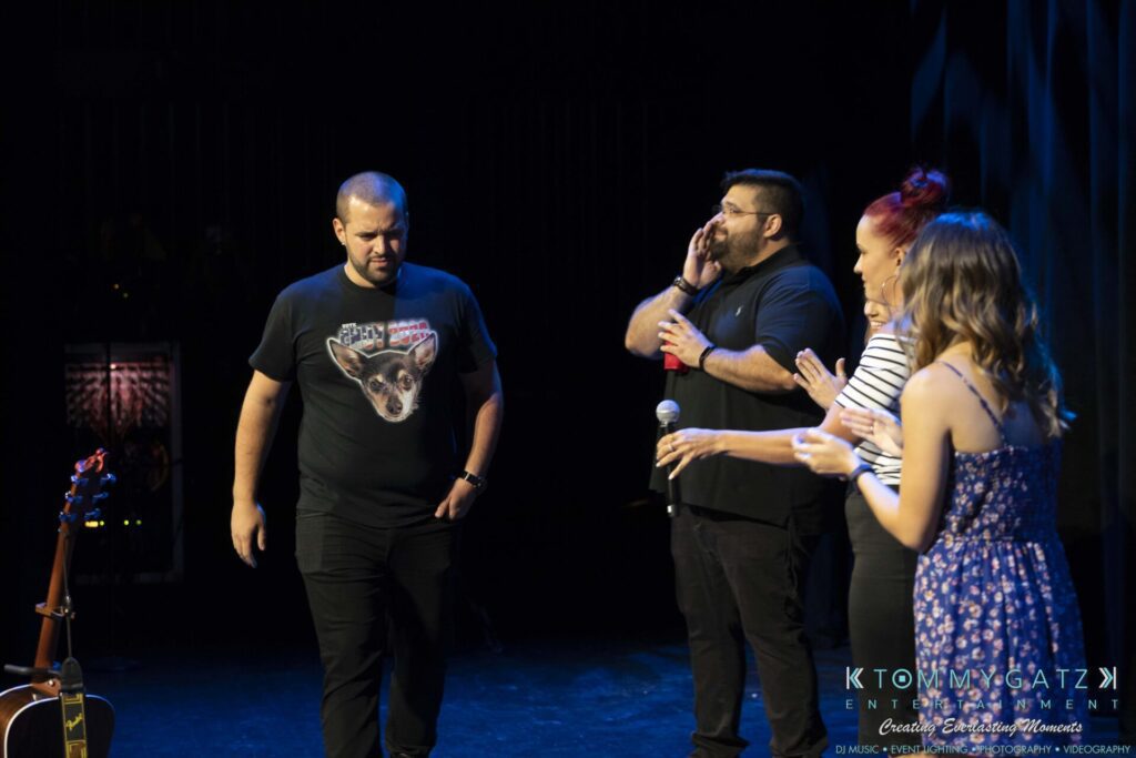 A group of four people on stage, with one gesturing and others looking on. A guitar is visible on the left. Stage lighting and equipment in the background.