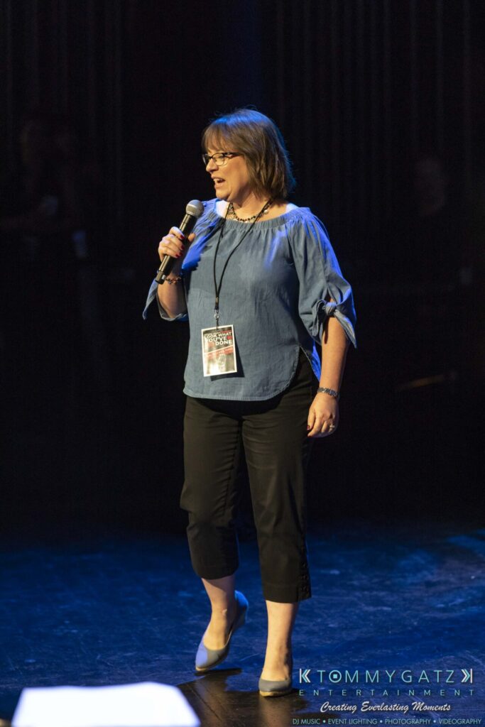 A woman in a blue blouse and black pants speaks into a microphone on stage.