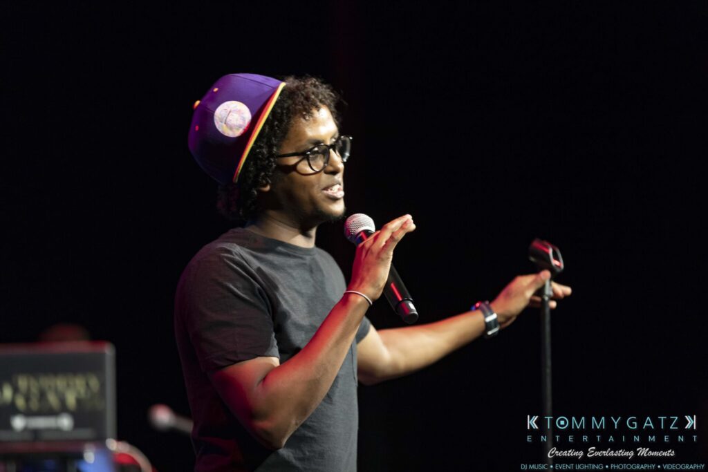 A person wearing a purple hat and black shirt holds a microphone on stage.