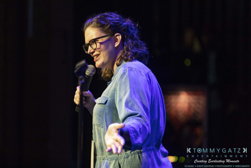 A person wearing glasses and a denim jacket is speaking into a microphone on stage, gesturing with one hand.