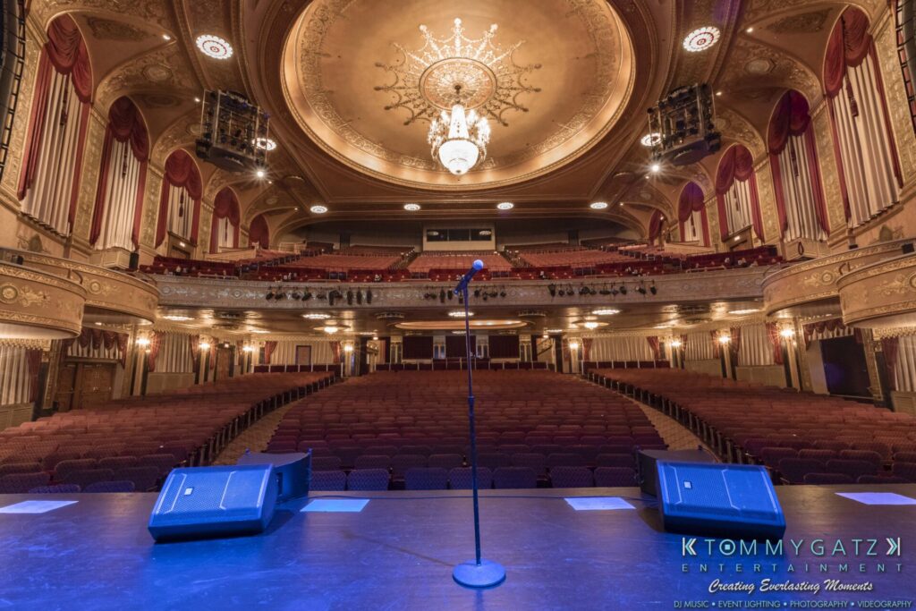 A grand theater view from the stage showing an empty audience, ornate ceiling, red curtains, and a microphone stand at the center with stage lights.