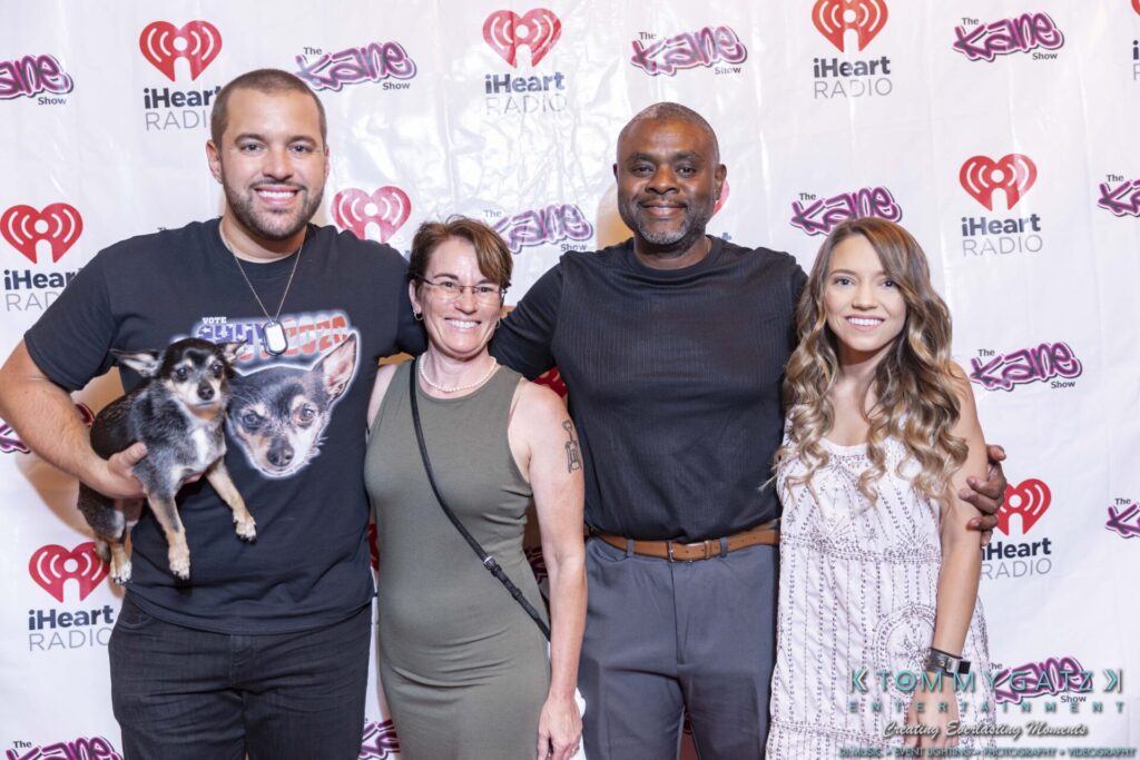 Four people smiling in front of a step and repeat banner featuring iHeartRadio and show logos. One person holds a small dog.