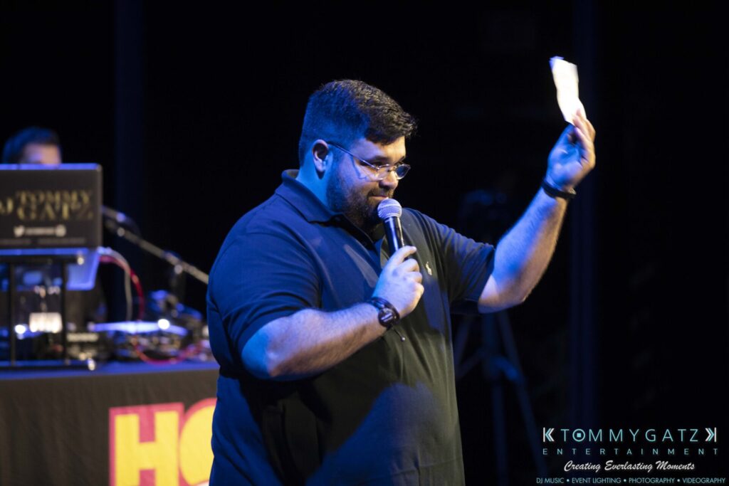 A person holding a microphone and a piece of paper, standing on stage. There's audio equipment in the background.