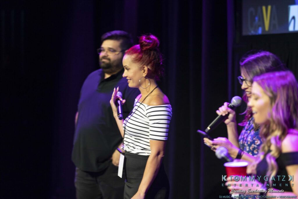 Three people are speaking on stage with microphones. A fourth person stands nearby. The background is dark, and a small logo is visible in the corner.