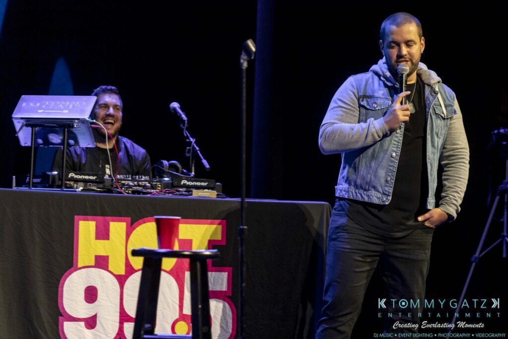 Two men on stage; one is sitting behind DJ equipment and laughing, the other is standing with a microphone, wearing a denim jacket. The background features the "Hot 99.5" logo and stage lighting.