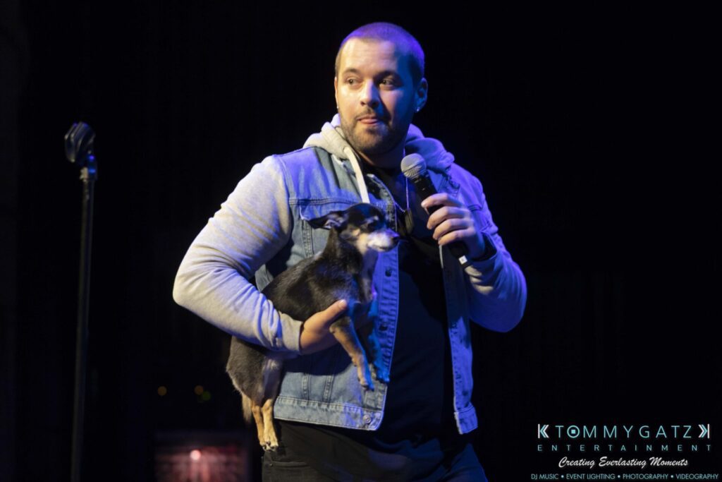A man holds a small dog in one arm while standing on stage with a microphone.