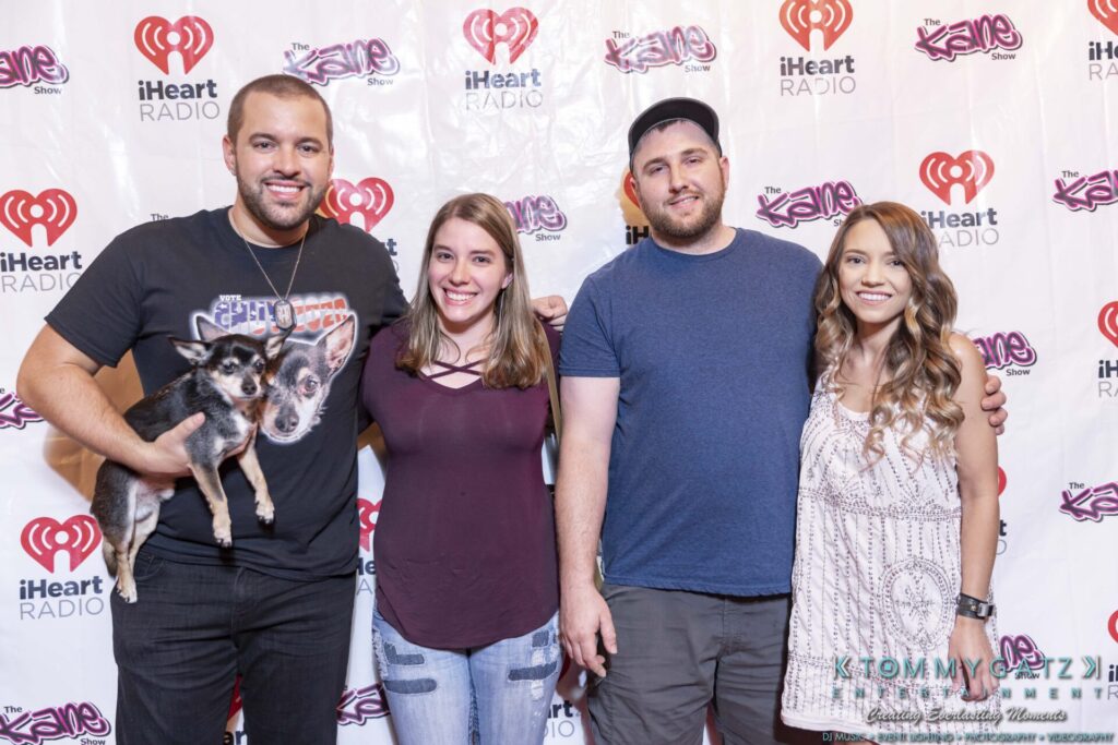 Four people stand in front of an event backdrop with iHeart Radio logos; one person holds a small dog.