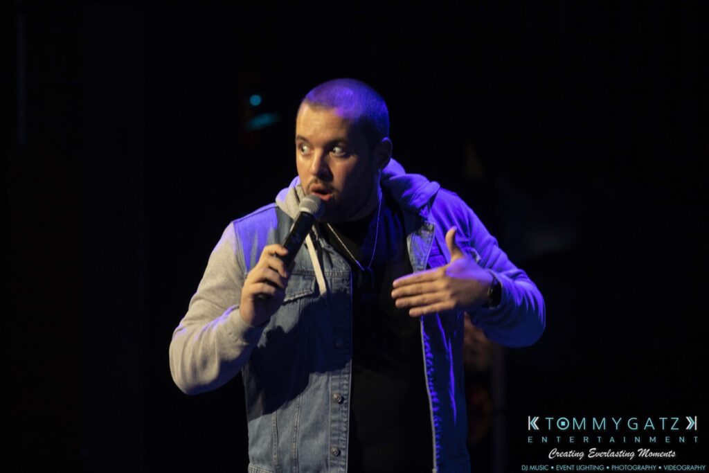 A person is speaking on stage with a microphone, wearing a denim jacket and hoodie. The background is dark.