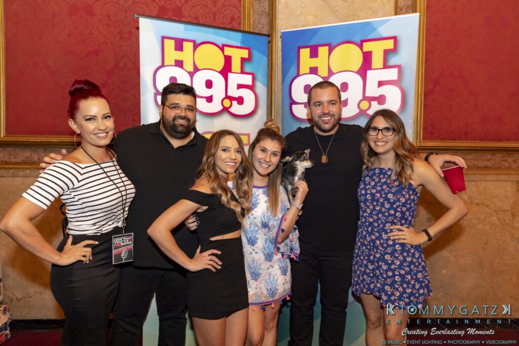 Group of six people smiling in front of Hot 99.5 banners, with one person holding a small dog.
