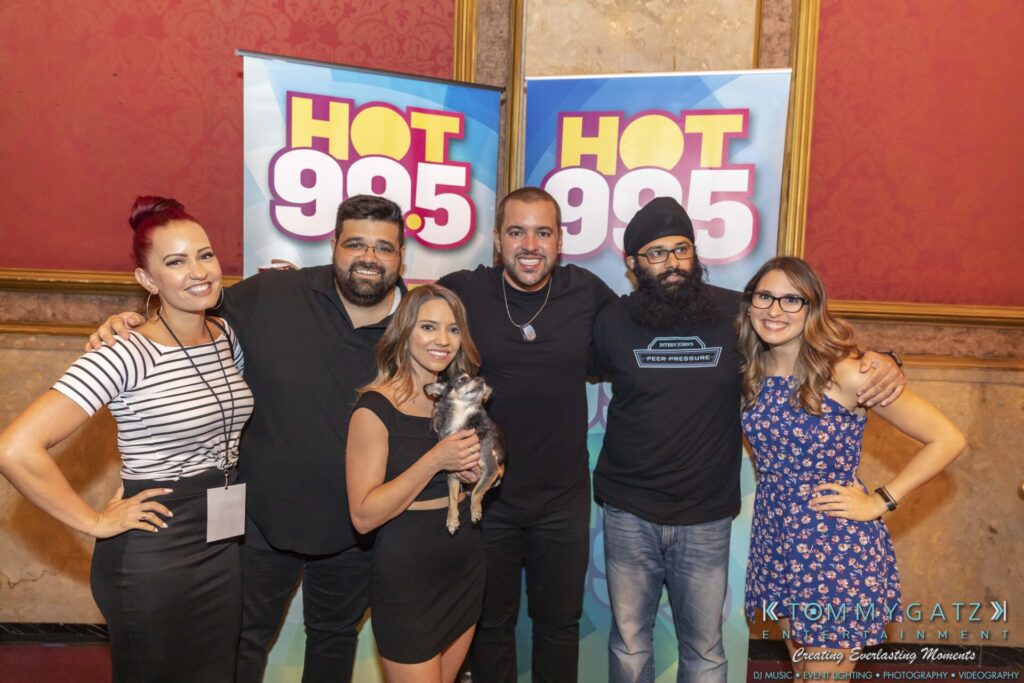 A group of six people posing in front of "HOT 99.5" banners; one person is holding a small dog.