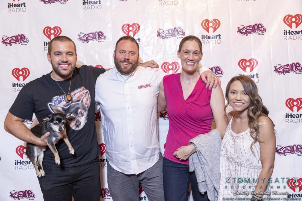 Four people posing in front of an iHeartRadio backdrop; one holds a small dog.