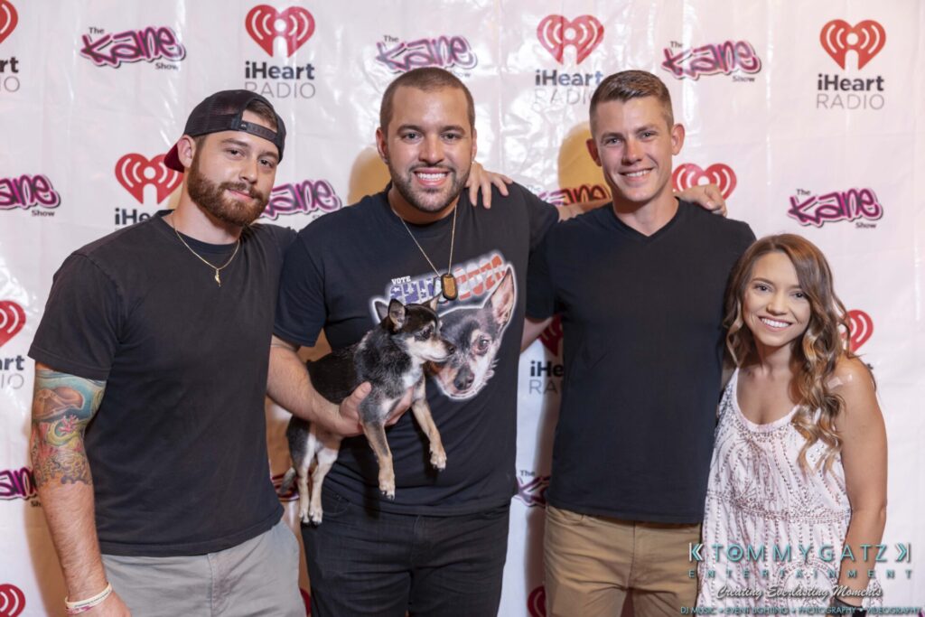 Four people and a dog pose for a group photo in front of an iHeart Radio and Kane Show backdrop.