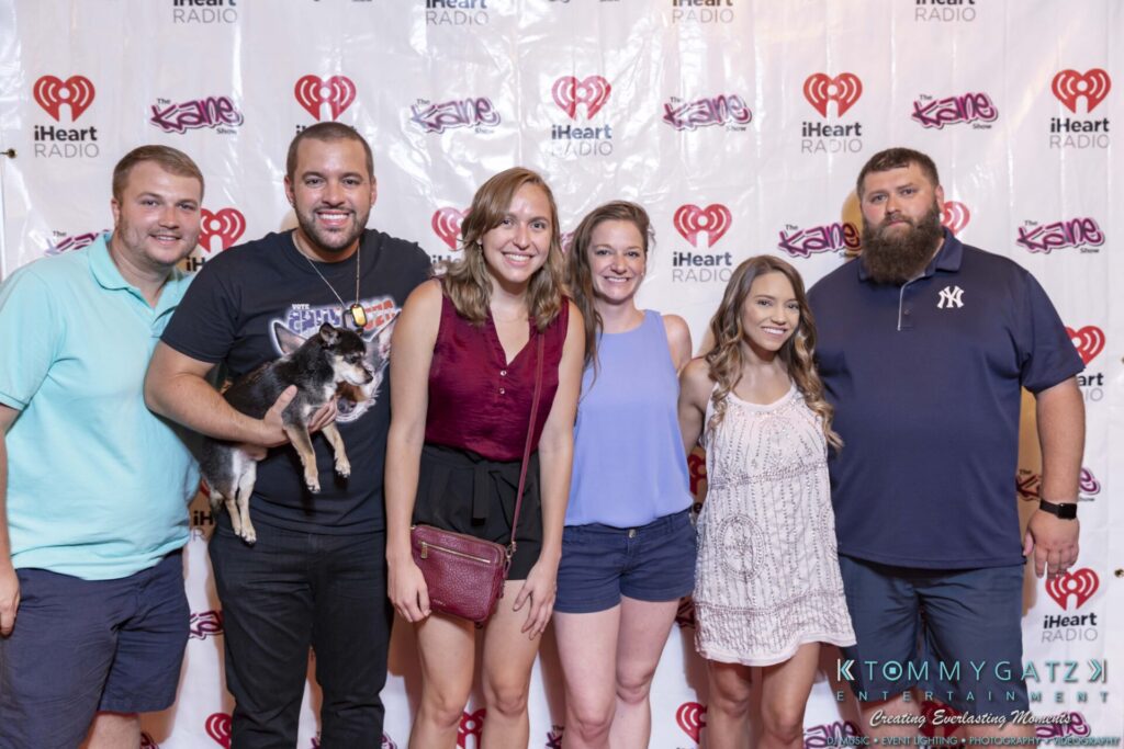 Group of six people smiling in front of an iHeartRadio backdrop; one person is holding a small dog.