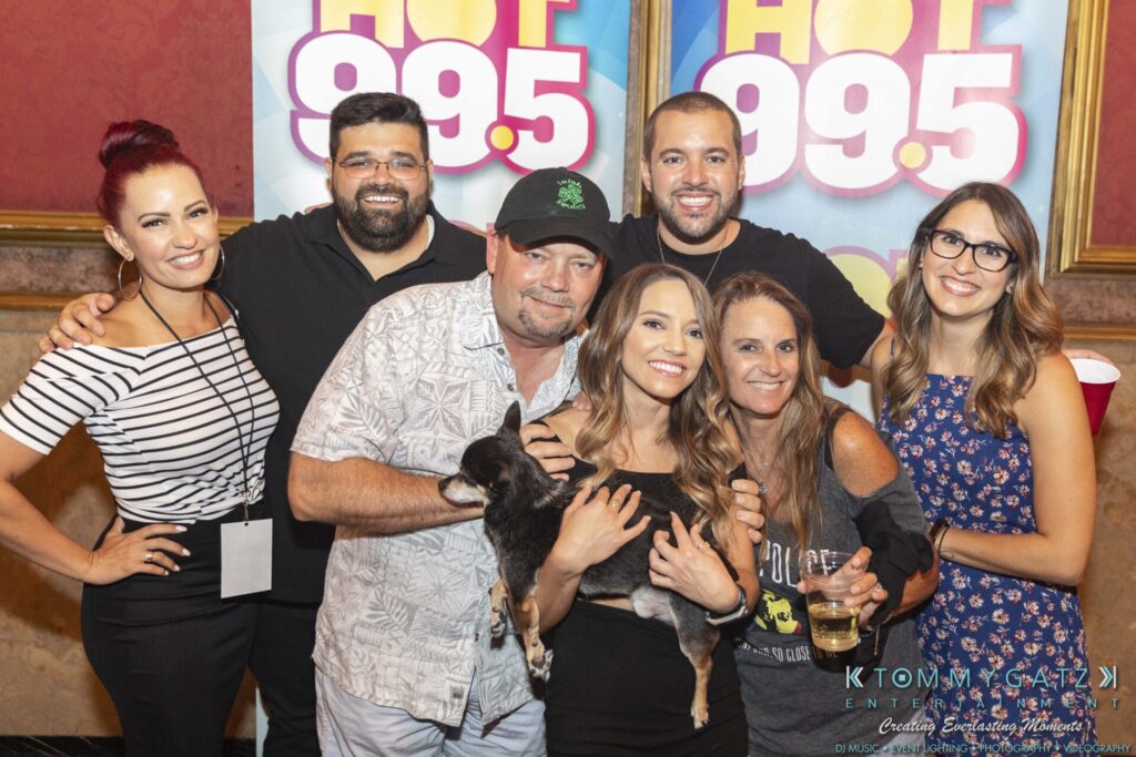 A group of seven people posing for a photo in front of "Hot 99.5" banners, with one woman holding a small dog.