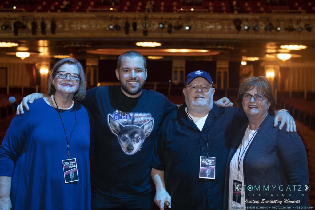 Four people stand together in a theater setting, wearing name badges and casual clothing, smiling at the camera.