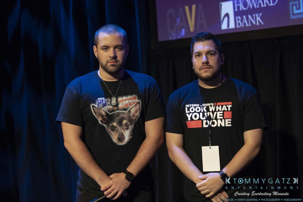 Two men standing on a stage under dim lighting, one wearing a T-shirt with a dog's face and the other with text, against a backdrop with logos.