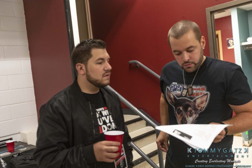 Two men on a staircase, one holding a red cup and the other reading a paper. A sign for "Tommy Gatz Entertainment" is visible.