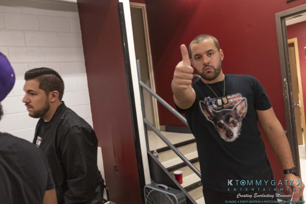 A man stands indoors giving a thumbs-up gesture. Another man is nearby. A staircase and a red wall are in the background.