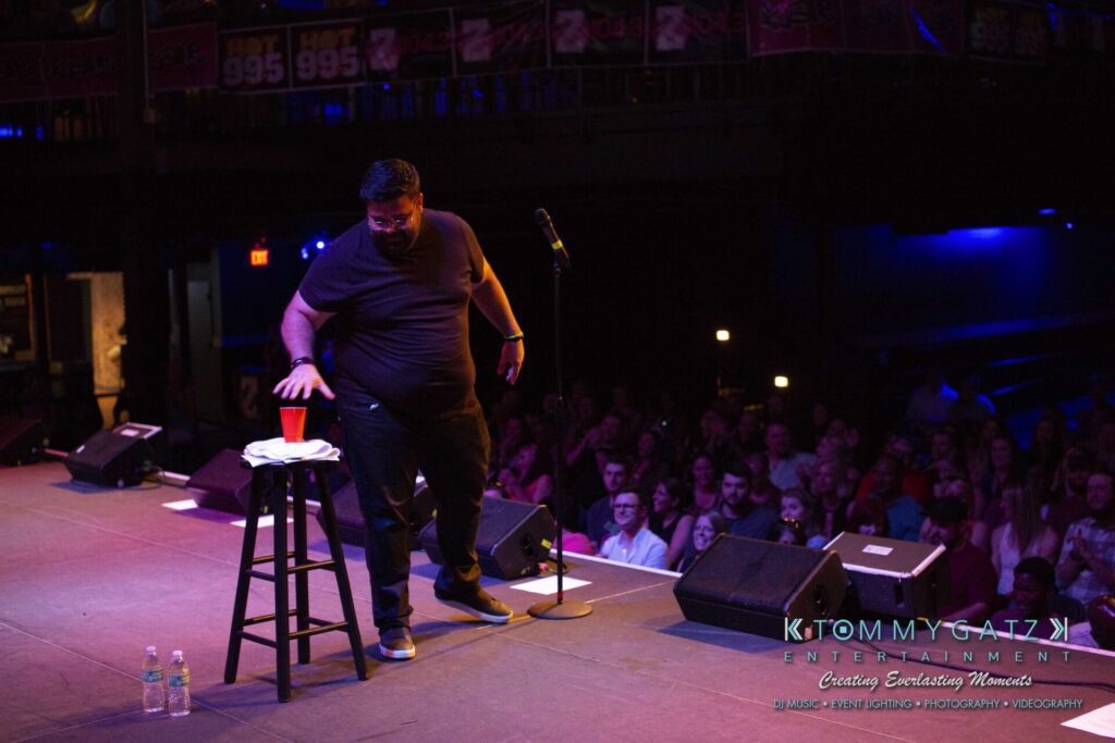 LWYD-Comedy-Tour-Baltimore-060118-FINALS-Low-Res-Watermark-101 - TOMMY GATZ ENTERTAINMENT A performer on stage reaches for a cup on a stool, with an audience seated in the background.