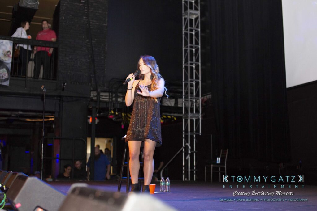 LWYD-Comedy-Tour-Baltimore-060118-FINALS-Low-Res-Watermark-110 - TOMMY GATZ ENTERTAINMENT A woman in a black dress is speaking into a microphone on a stage with speakers and a screen in the background.