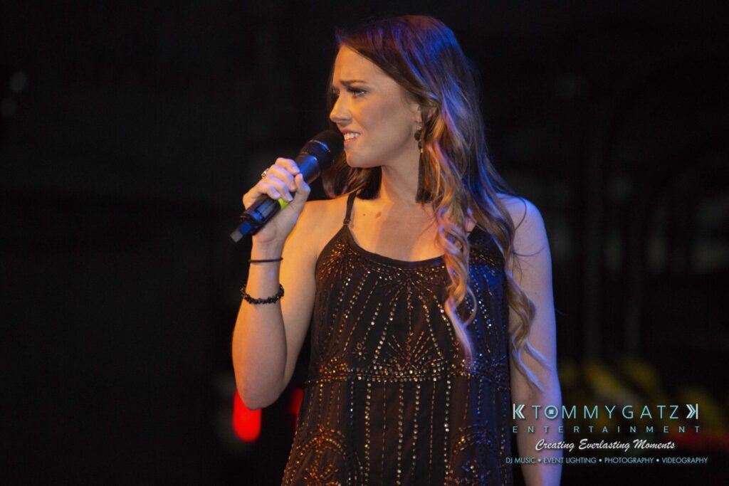 LWYD-Comedy-Tour-Baltimore-060118-FINALS-Low-Res-Watermark-116 - TOMMY GATZ ENTERTAINMENT A woman holding a microphone, wearing a black patterned dress, stands on stage looking to her left.