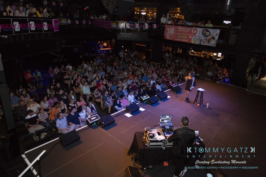 LWYD-Comedy-Tour-Baltimore-060118-FINALS-Low-Res-Watermark-122 - TOMMY GATZ ENTERTAINMENT A comedian performs on stage in front of a large seated audience at an indoor venue. Audio and lighting equipment are visible in the foreground.