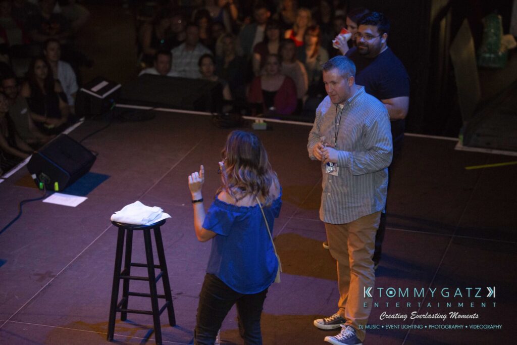 LWYD-Comedy-Tour-Baltimore-060118-FINALS-Low-Res-Watermark-125 - TOMMY GATZ ENTERTAINMENT A woman on stage holds a microphone facing a seated audience, with two men standing nearby. A black stool with a white cloth is on stage, alongside a sign for Tommy Gatz Entertainment.