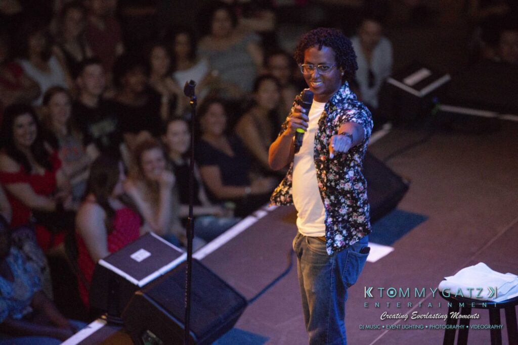 LWYD-Comedy-Tour-Baltimore-060118-FINALS-Low-Res-Watermark-128 - TOMMY GATZ ENTERTAINMENT A person on stage holds a microphone, addressing a smiling audience.