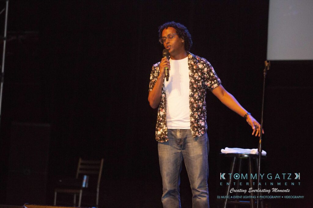 LWYD-Comedy-Tour-Baltimore-060118-FINALS-Low-Res-Watermark-129 - TOMMY GATZ ENTERTAINMENT A person in a floral shirt performs stand-up comedy on stage with a microphone, next to a stool.