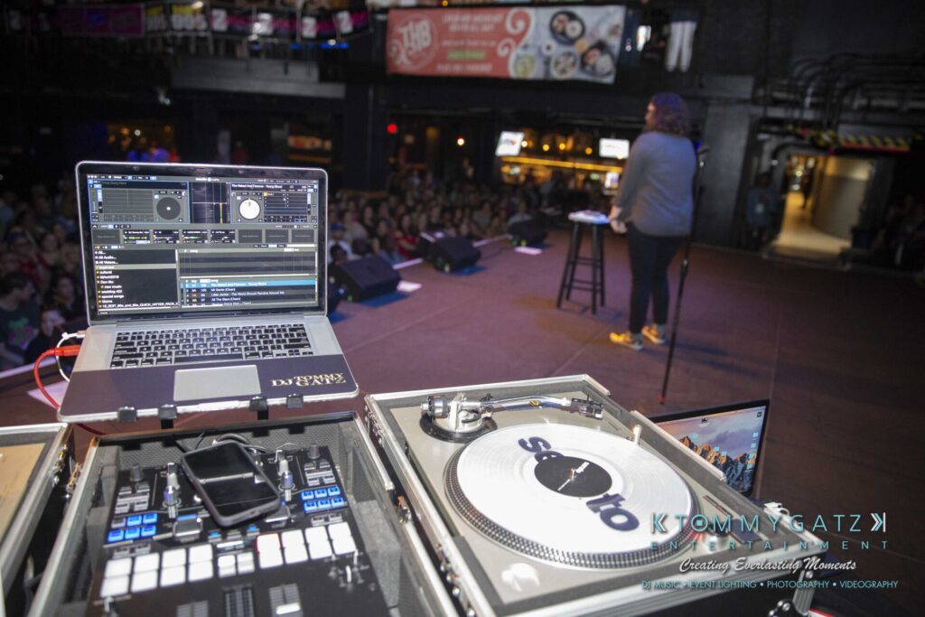 LWYD-Comedy-Tour-Baltimore-060118-FINALS-Low-Res-Watermark-156 - TOMMY GATZ ENTERTAINMENT DJ setup with a laptop and turntable in the foreground. A speaker stands on stage addressing an audience in a dimly lit venue.