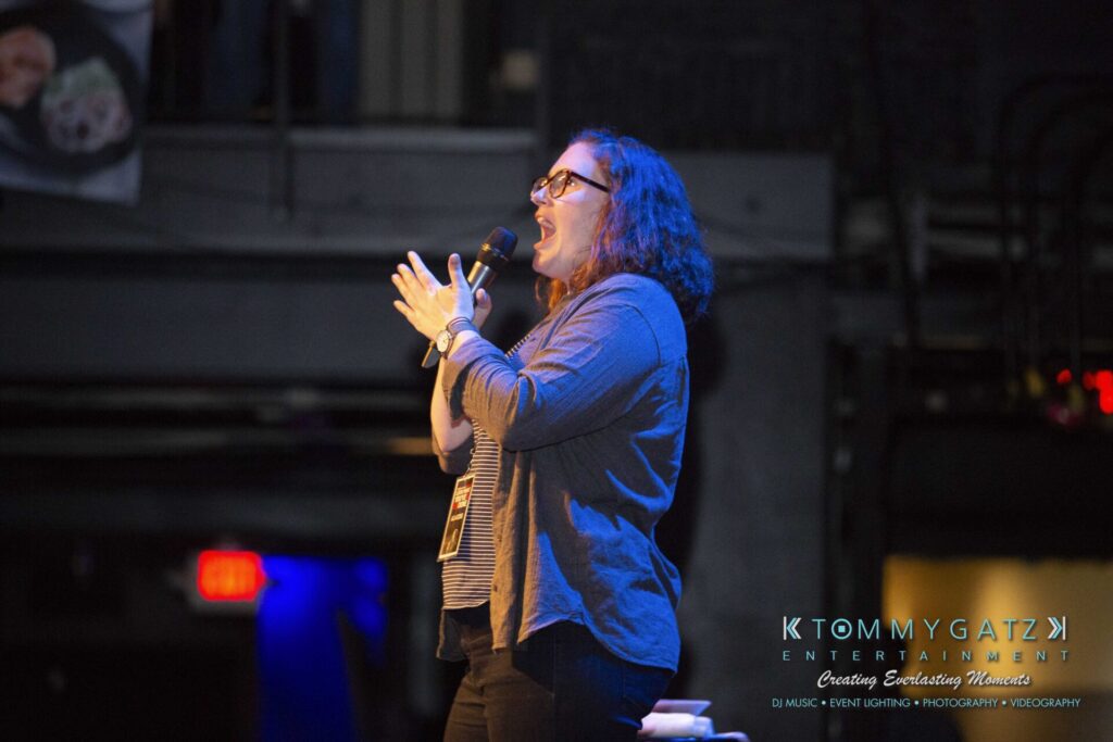 LWYD-Comedy-Tour-Baltimore-060118-FINALS-Low-Res-Watermark-169 - TOMMY GATZ ENTERTAINMENT A woman with glasses speaks into a microphone on stage. She is wearing a gray cardigan and dark pants. The backdrop shows a logo for Tommy Gatz Entertainment.